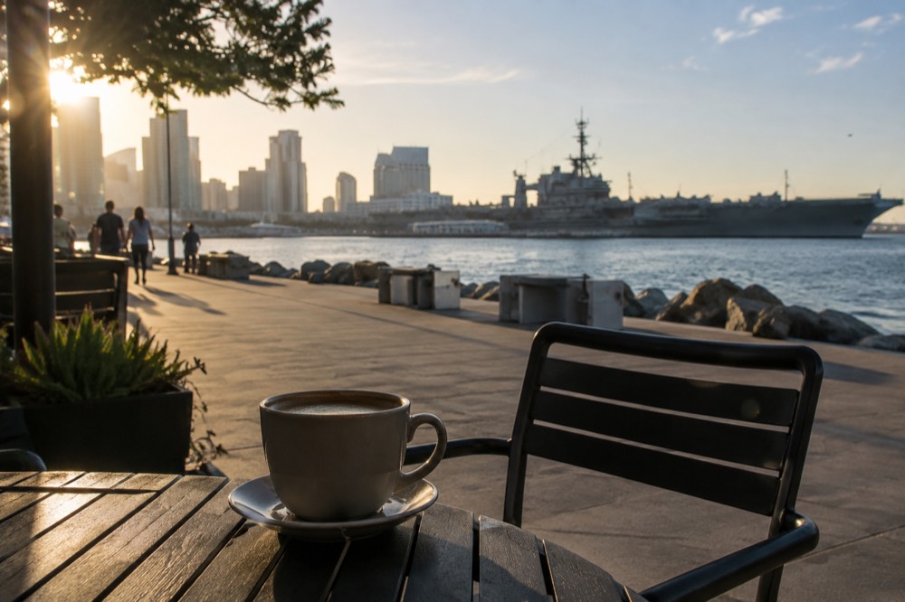 A coffee cup on an outdoor table beside a waterfront promenade with ships in the background