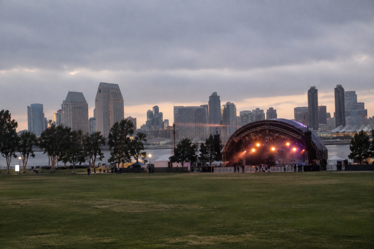 An outdoor concert stage at dusk with stage lights glowing over an empty green field