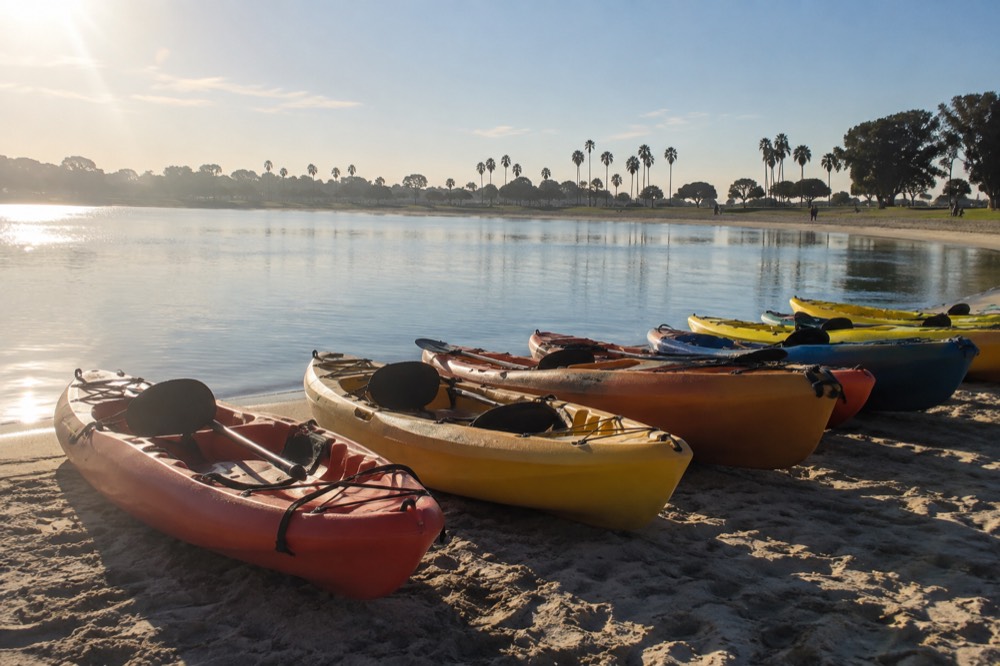 Kayaks lined up on a sandy beach beside still bay water in the morning