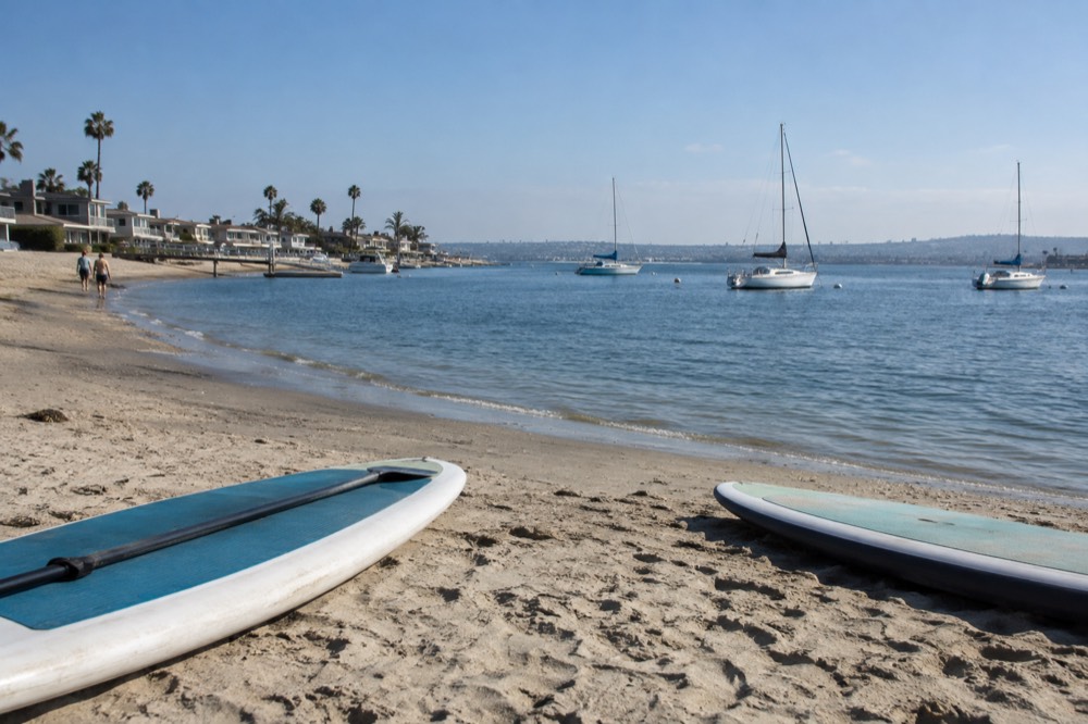 A calm bay with paddleboards and small sailboats floating near a sandy shore