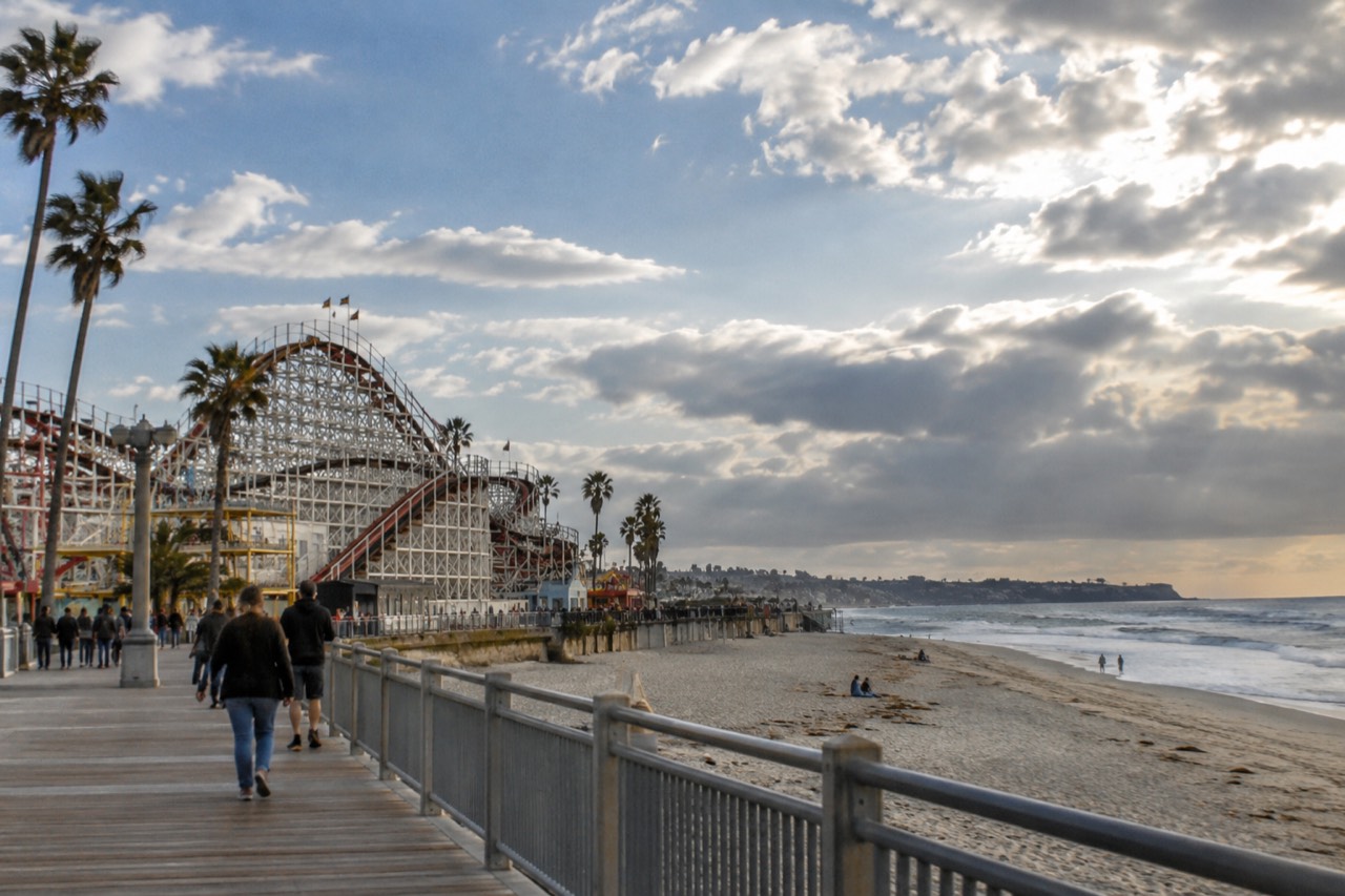 A seaside amusement park boardwalk with a wooden roller coaster rising above the beach