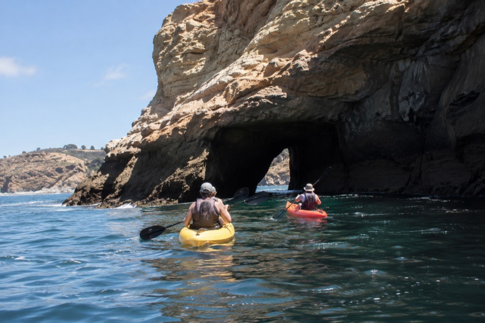 Two sit-on-top kayaks paddling close to a sandstone cliff with a cave arch