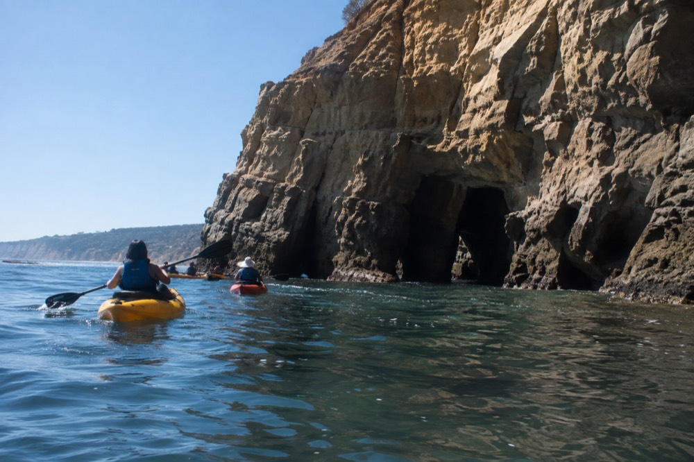 Kayakers paddling along the cliff base toward the sea caves on a clear morning