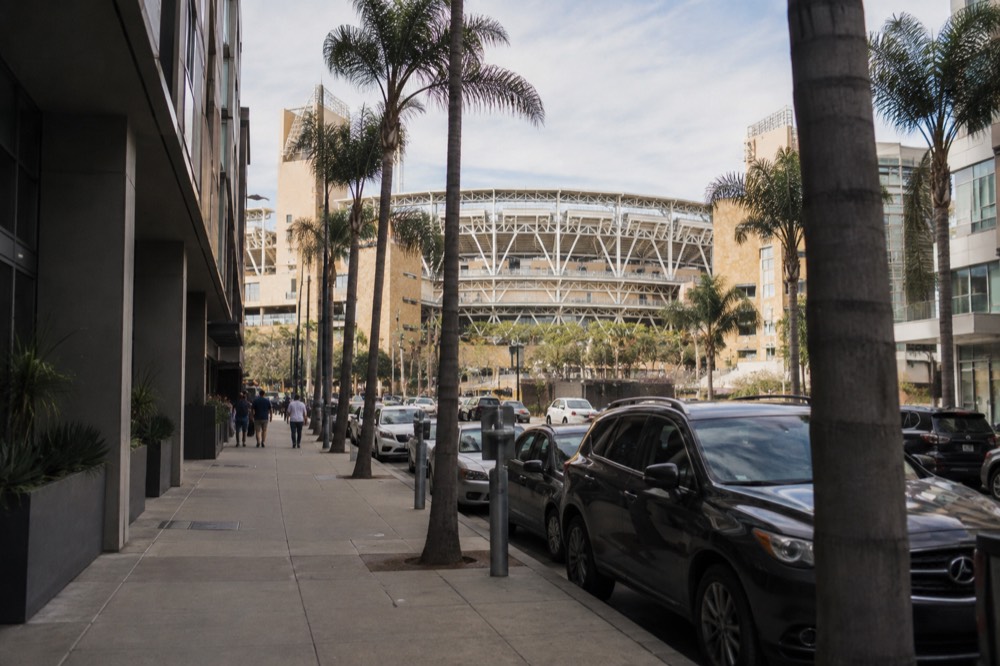 A downtown street with palm trees, parked cars and a ballpark entrance in the distance
