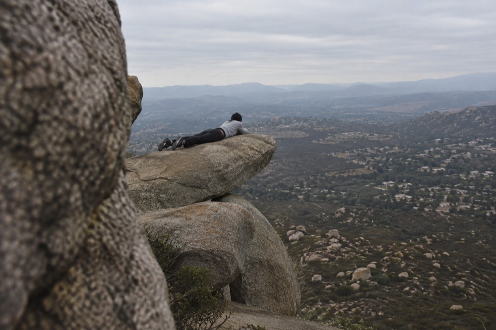 A person lying flat on a thin rock ledge with a wide valley far below