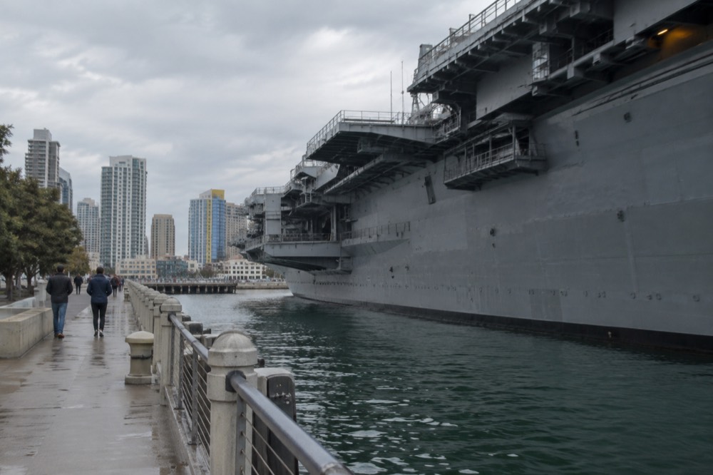 An aircraft carrier moored at a waterfront dock with a walkway and city buildings behind it