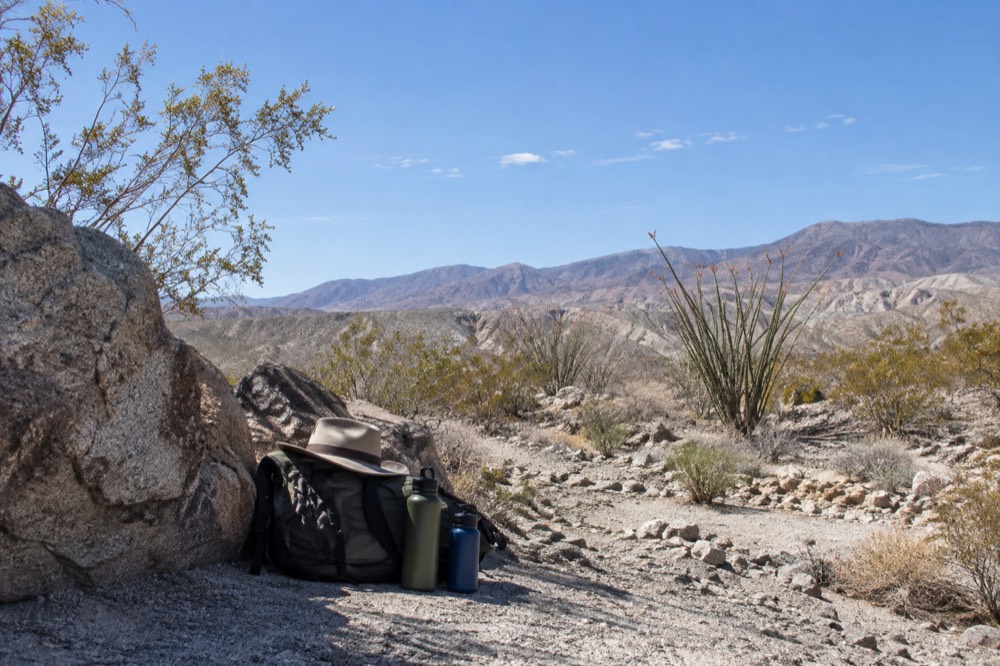 Backpack, wide-brim hat and water bottles arranged on a rock in desert shade