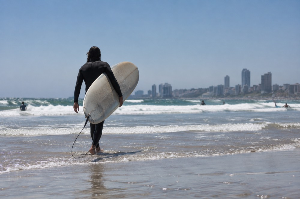 Surfer walking a longboard across wet sand toward small breaking waves
