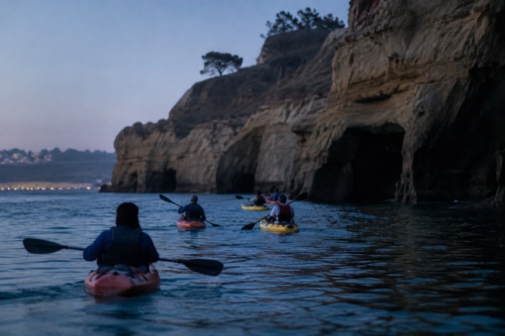 A group of kayaks paddling over calm turquoise water toward sea cliffs