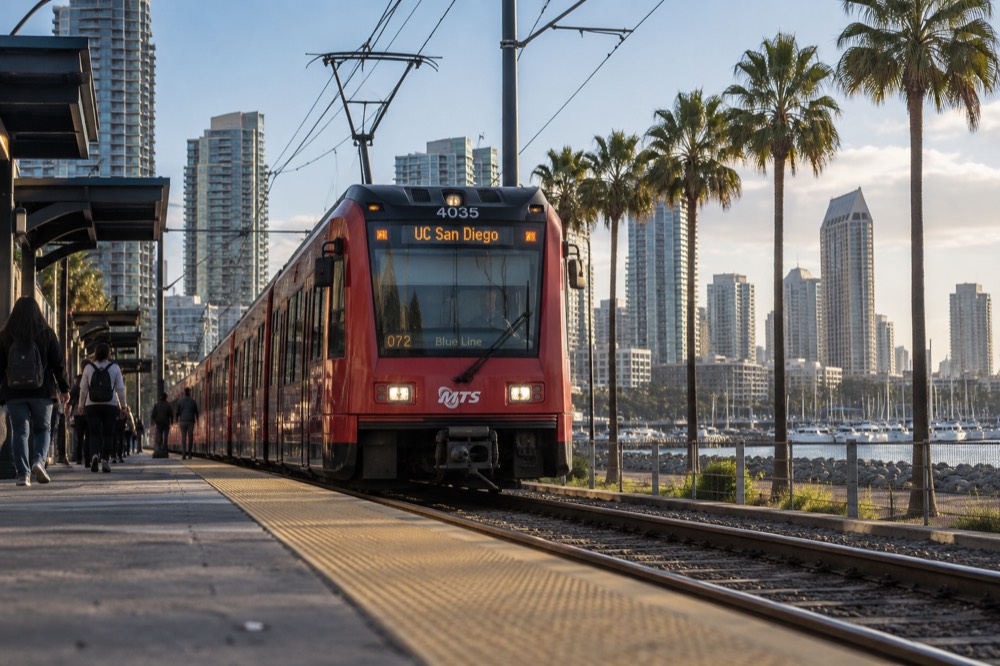 A red trolley car approaching a station platform beside palm trees and a bay