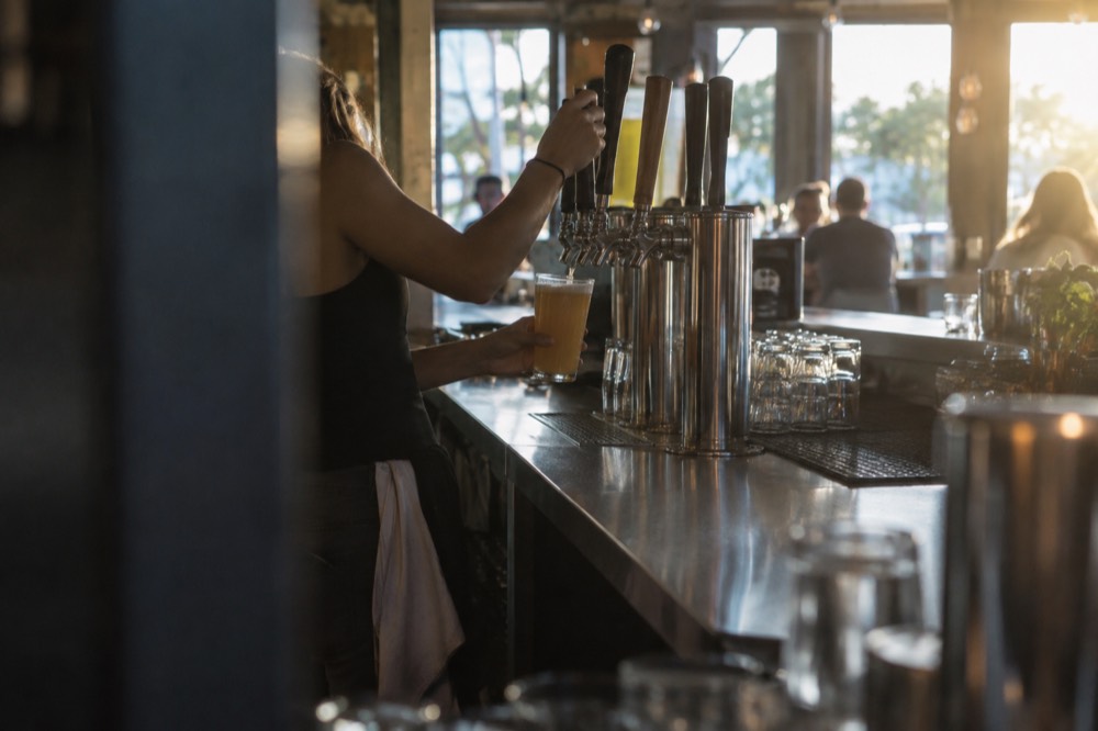 A bartender pouring beer from a tap handle at a long stainless steel bar