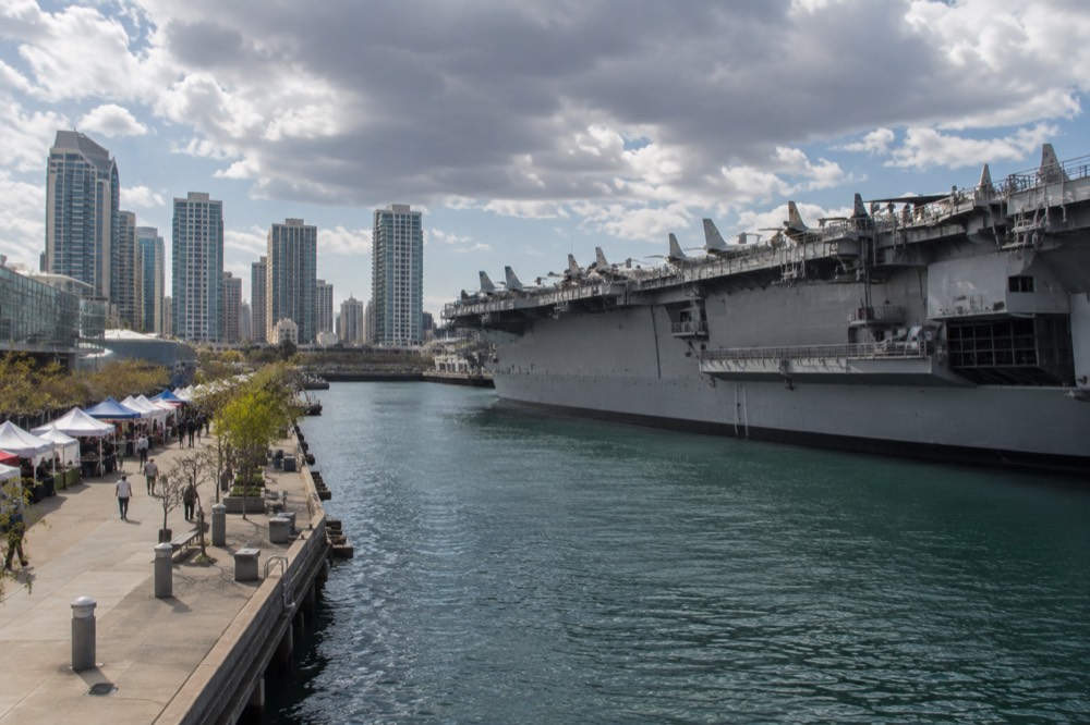 A harbor view with a gray aircraft carrier and downtown buildings beyond a market street