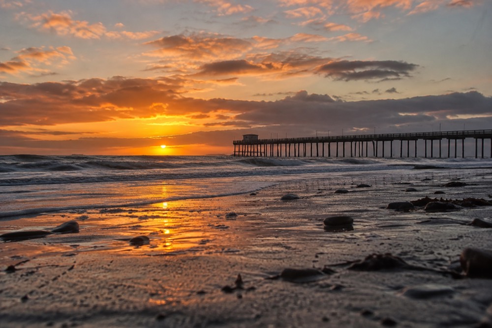 Wide flat beach at dusk with a long wooden silhouette against orange sky