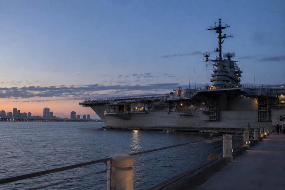 A gray aircraft carrier moored at a downtown waterfront pier
