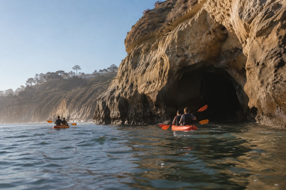 Two-person kayaks paddling along a sandstone cliff toward a sea cave