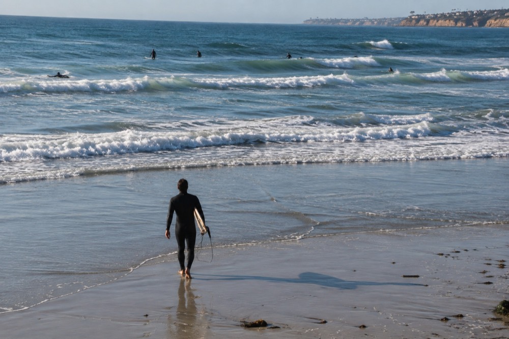 Surfer in a full wetsuit walking across wet sand toward small waves