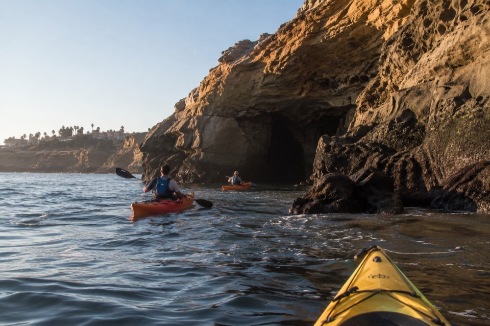 Kayaks paddling along rocky cliffs toward a sea cave on a clear morning