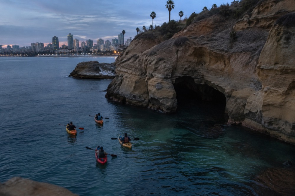 Kayaks paddling over clear green water toward a sea cave opening in a sandstone cliff